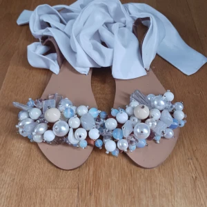 Elina Linardaki handgjorda tofflor - Unika sandaler med beige sula och vita band att knyta runt vristen. Framtill dekorerade med stora pärlor och stenar i vitt, blått och silver. Perfekta för dig som vill sticka ut med detaljerade accessoarer. Storlek 38. Helt nytt skick aldrig använda. Nypris 2500kr pris går att diskutera.