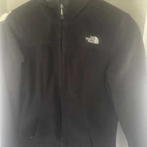 Svart fleecejacka från The North Face - Säljer en svart fleecejacka från The North Face med dragkedja framtill och hög krage. Jackan har lång ärm och är tillverkad i mjuk fleece som håller dig varm. Perfekt för kyliga dagar och enkel att matcha med andra plagg. den är i ett bra skick och storlek s men den va för liten för mig så skulle säga att den passar mer xs 
