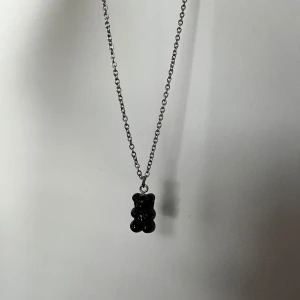 Halsband med svart nallebjörn-berlock - Snyggt halsband med en svart nallebjörn-berlock i plast. Kedjan är tunn och silverfärgad, vilket ger ett stilrent och lekfullt intryck. Perfekt för dig som vill ha något unikt och trendigt i smyckeslådan.