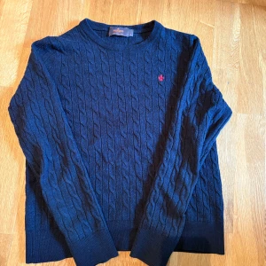Mörkblå kabelstickad tröja från Morris - Säljer en mörkblå kabelstickad tröja från Morris i 100% merinoull. Tröjan har rund halsringning, lång ärm och en röd broderad logga på bröstet. Perfekt för lager-på-lager och snygg till jeans eller kjol.
