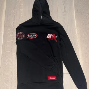 Svart hoodie bull från Mercier med patches - Svart hoodie från Mercier i storlek XS med röda och vita broderade patches och loggor på bröstet, ärmen och ryggen. Tröjan har huva, långa ärmar och en klassisk loose passform. Perfekt för dig som gillar streetwear och snygga detaljer. Har ej använt den.
