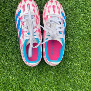 Adidas Predator fotbollsskor vit/blå/rosa - Säljer ett par Adidas Predator fotbollsskor i vitt med blå och rosa detaljer. Skorna har snörning, mönstrad ovandel och dobbar i blått och rosa för bra grepp på planen. Insidan är fodrad i blått och rosa för extra stilpoäng. Perfekta för dig som vill sticka ut på fotbollsplanen.