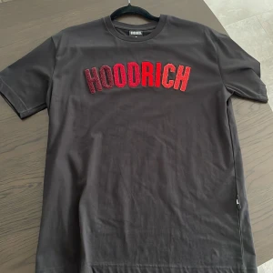 Svart HOODRICH t-shirt med röd text - Svart t-shirt från HOODRICH i bomull med stor broderad text i rött och vinrött över bröstet. Klassisk rund halsringning och korta ärmar. Perfekt för en streetwear-look och enkel att matcha med jeans eller shorts.
