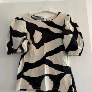 Svartvit blus med puffärm från H&M - Cool blus från H&M med svartvitt abstrakt mönster och puffiga korta ärmar. Rund halsringning och rak passform. Materialet är mjuk bomull som känns skönt mot huden. Perfekt för dig som gillar statement-plagg med unik design.