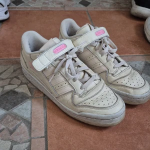 Adidas Forum Low i beige och rosa - Snygga Adidas Forum Low sneakers i beige skinn med rosa detaljer och klassiska tre ränder på sidorna. Skorna har kardborreband som går att ta bort. De är rätt så använda så lite smutsiga, nopper på insidan och inte bästa skick. Med lite jobb kan de bli riktigt snygga igen. Pris kan absolut diskuteras.