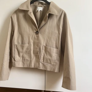 Beige kappa från H&M, storlek S - Snygg beige kappa från H&M i polyester med klassisk krage, stora framfickor och knäppning med knappar framtill.  Den är helt ny och har inte fått någon användning, ny utan prislapp!