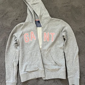 Grå hoodie från GANT med dragkedja - Grå hoodie från GANT med rosa bokstäver framtill. Tröjan har huva, dragkedja och två fickor på magen. Tillverkad i mjuk bomullsmix och har långa ärmar med ribbade muddar. Perfekt för en chill och avslappnad stil.