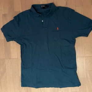 Blå Polo Ralph Lauren piké🤩 - Snygg blå piké från Polo Ralph Lauren med klassisk krage och två knappar framtill. Ikoniska orangea loggan broderad på bröstet. Tillverkad i mjuk bomull som känns skön mot huden. Perfekt för dig som gillar stilren och enkel design.