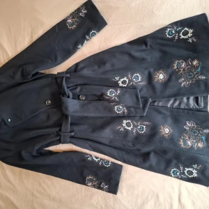 Broderad svart kappa från Fransa Storlel 160-170 - Svart kappa från Fransa med broderade blommor i blått, vitt och beige. Knytbälte i midjan, knappar framtill och långärmad modell. Perfekt för dig som vill ha en unik och snygg jacka med coola detaljer.