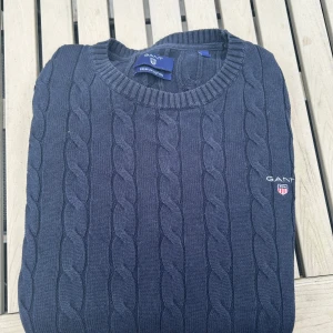 Mörkblå stickad tröja från GANT - Säljer en stilren mörkblå stickad tröja från GANT i premium bomull. Tröjan har ett klassiskt kabelstickat mönster och rund halsringning. Perfekt för kyligare dagar.