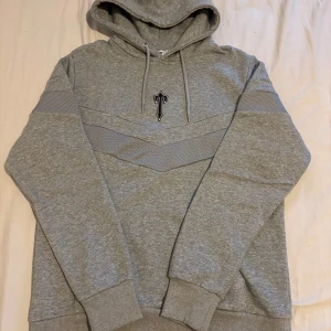 Grå hoodie från Trapstar med T-logga - Säljer en grå hoodie från Trapstar i storlek L. Tröjan har en stor huva med snörning, långärmade ärmar och ett snyggt T-emblem broderat på bröstet. Materialet är mjukt och skönt, med mesh-detaljer i ett V-mönster över bröst och rygg. Perfekt för chill eller streetwear. Pris kan diskuteras 