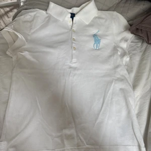 Vit pikétröja från Ralph Lauren - Vit pikétröja från Ralph Lauren med klassisk krage, korta ärmar och knappar framtill. Stor ljusblå broderad logga på bröstet. Tillverkad i bomull för en skön och sportig känsla. Perfekt för en clean och stilren look. Storlek L (12-14år).