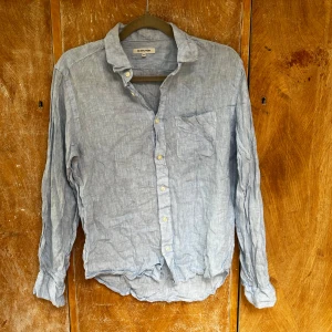 Ljusblå linneskjorta Global Work M - Snygg ljusblå skjorta från Global Work i luftig linne. Klassisk krage, bröstficka och knappar framtill. Perfekt för varmare dagar och enkel att styla till jeans eller shorts. Långärmad och avslappnad passform.