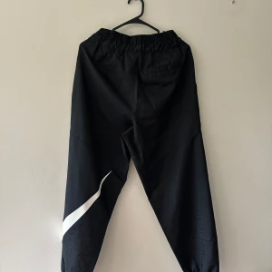 Svarta Nike mjukisbyxor med logga - Svarta mjukisbyxor från Nike med vit swoosh-logga på ena benet. Byxorna har resår i midjan med snörning och elastiska benslut. Perfekta för chill eller träning, tillverkade i mjukt syntetmaterial för skön passform.