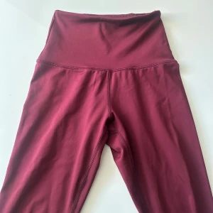 Vinröda högmidjade träningsbyxor  - Snygga vinröda träningstights med hög midja och tight passform. Perfekta för träning eller chill, tillverkade i mjukt och stretchigt syntetmaterial som sitter skönt mot kroppen. Stilrena utan detaljer och med långa ben för en clean look.