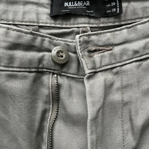 Gråa raka byxor från Pull&Bear - Snygga gråa byxor från Pull&Bear med lite olivgrön underton. Rakt snitt och normal passform, perfekta för en clean och stilren look. Byxorna har dragkedja och knapp framtill samt hällor för bälte. Tillverkade i bomullsmix för skön känsla samt medel till högmidjade. Vid frågor hör av dig!