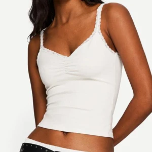 Vit  linnetopp med spetskant - Supersöt vit linnetopp med smala axelband och spetskant längs ringningen. Toppen har en lätt rynkning vid bysten och är croppad i modellen. Perfekt att styla med jeans eller kjol för en fräsch look.💕