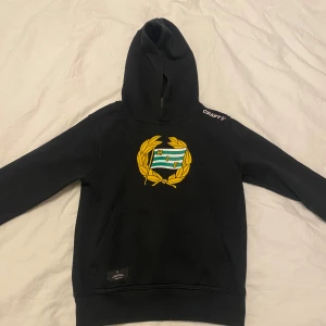 Svart Hammarby hoodie från Craft - Svart hoodie från Craft med Hammarby IF:s klassiska emblem i gult och grönt på bröstet. Tröjan har huva, magficka och långa ärmar. 