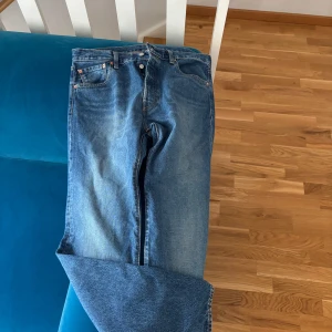 Levi's 501 blå jeans W34 L32 - Klassiska Levi's 501 jeans i blå denim, rak passform och knappgylf. Snyggt slitna detaljer och ikonisk läderpatch bak. Premiumkvalitet med fem fickor och orange sömmar. Perfekta för dig som gillar tidlös stil. De är lite mer åt baggy sidan i sin passform! Använda cirka 3 gånger.