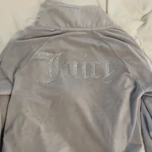 Ljusgrå kofta från Juicy Couture - Supermjuk ljusgrå kofta från Juicy Couture med glittrande Juicy-text på ryggen och JC-detalj fram. Jackan har dragkedja med rund metalltag, ribbade muddar och två fickor framtill. Endast använd en gång