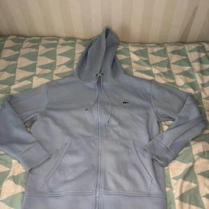 Ljusblå hoodie från Lacoste med dragkedja - Säljer en ljusblå hoodie från Lacoste i klassisk passform. Tröjan har huva med snörning, dragkedja framtill och två fickor. Ikoniska Lacoste-loggan på bröstet. Perfekt för chill dagar och snygg till jeans eller joggers.