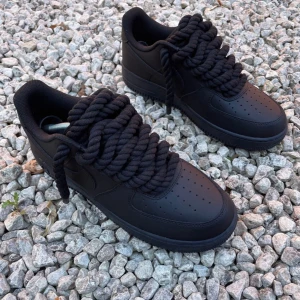 Nike Air Force 1 svart med chunky snören - Svarta Nike Air Force 1 sneakers med riktigt chunky rep-liknande snören som ger en unik och fet look. Skorna är i helsvart skinn med perforerad tå och klassisk siluett. Flera extra snören i olika färger som beige, blå, vit, röd och rosa medföljer för att variera stilen.
