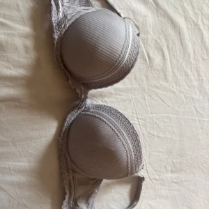 Ljusgrå ribbad bh med spetsdetaljer - Snygg ljusgrå bh med ribbat tyg och fina spetsdetaljer längs kanterna. Kuporna är vadderade och insidan är slät och mjuk. Axelbanden är justerbara för bästa passform. Perfekt för dig som gillar en stilren och feminin look.