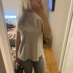 Beige ärmlös polotopp från Zara - Säljer en stilren beige polotopp från Zara. Toppen är ärmlös med hög, ribbad polokrage och har en rak, loose passform. Perfekt att styla med jeans eller kjol för en clean look. Materialet är mjukt och känns skönt mot huden.