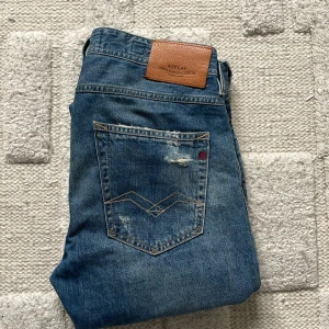 Blå slitna jeans från Replay Willbi - Säljer ett par blå Replay Willbi jeans med slim fit och snygga slitningar och hål på både fram- och baksida. Klassisk femficksmodell med läderpatch bak och detaljerade sömmar på bakfickan. Perfekta för dig som gillar en avslappnad och trendig look.