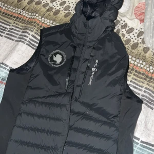 Svart dunväst Sail Racing med huva - Svart dunväst från Sail Racing, modell Antarctica Hybrid Vest. Västen har en vadderad, quiltad design med huva och dragkedja framtill. Snygg logga på bröstet och detaljer på huvan. Tillverkad i nylon och polyester med dunfyllning för extra värme.