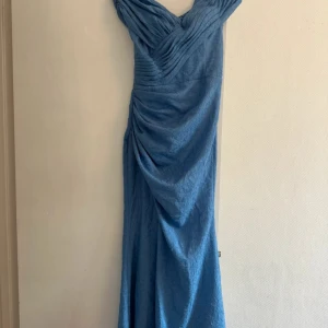 Lång blå klänning från VIP Girl - Säljer en lång blå klänning från VIP Girl i storlek S. Klänningen har snygga draperingar, offshoulder-skärning och snörning i ryggen. Materialet känns mjukt och lätt, perfekt för dig som vill sticka ut med en elegant look.