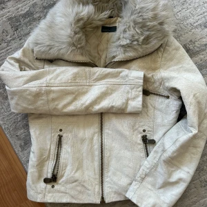 Beige pälsjacka från Rock'n Blue - Säljer en snygg beige pälsjacka från Rock'n Blue med stor fluffig fuskpälskrage. Jackan har dragkedjor på fickorna och framtill, samt quiltat foder på insidan. Perfekt för dig som vill ha en varm och trendig look i vinter.