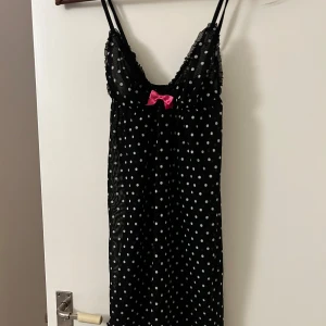 Svart prickig nattklänning med rosett - Säljer en svart natt kortklänning med vita prickar och en söt rosa rosett framtill. Klänningen har tunna axelband.