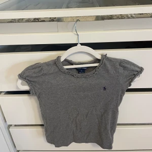 Ralph Lauren T-shirt kids - Supersöt grå t-shirt från Ralph Lauren kids med korta puffärmar, volangdetalj vid halsringningen och klassisk logga broderad på bröstet . Storlek 5 år (110-116)