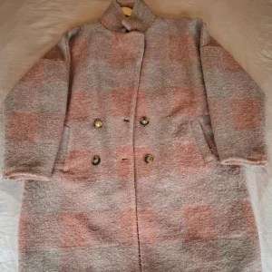 Rutig kappa från UNIQUE i ullmix stl M-L - Säljer en oversized rutig kappa från UNIQUE i grått och beige med inslag av rosa. Kappan har dubbelknäppning med stora knappar, hög krage och två fickor framtill. Materialet är en mjuk ullmix som känns både varm och stilren. Perfekt för kalla dagar! Storlek M-L