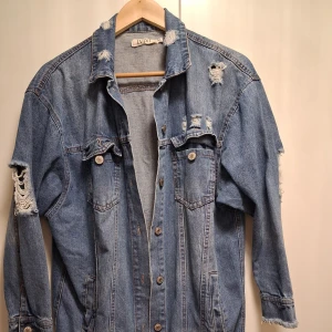 Oversized jeansjacka från IVIVI - Snygg oversized jeansjacka från IVIVI i klassisk blå denim. Jackan har slitna detaljer på axlar och ärmar, knappar framtill och två bröstfickor med lock. Perfekt för en chill och trendig look.