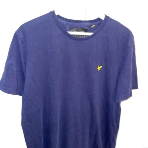 Marinblå t-shirt från Lyle & Scott - Snygg marinblå t-shirt från Lyle & Scott med klassisk rund hals och gul broderad logga på bröstet. Tillverkad i mjuk bomull och har en normal passform. Perfekt för en clean och stilren look.