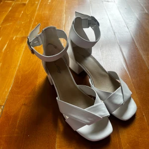 Vita klackar (Storlek 37)  - Snygga vita klackar från Alley med breda korsade remmar framtill och justerbart spänne runt ankeln. Skorna har en stabil låg klack och öppen tå, perfekta för sommarens alla tillfällen. Tillverkade i syntetmaterial med mjuk innersula.