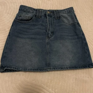 Blå jeanskjol med fickor - Säljer en klassisk blå jeanskjol med fickor och hällor för bälte. Kjolen har en rak, kort passform och stängs med knapp och dragkedja framtill. Perfekt för dig som gillar enkel och denimstil.