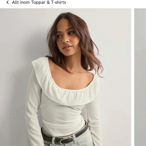 Vit topp med volang - Säljer en vit topp med bred volang runt halsringningen och långa ärmar. Toppen har en ribbad struktur och sitter snyggt med en figurnära passform. Perfekt för dig som gillar romantiska detaljer och stilren look.