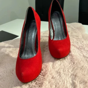 Röda mocka pumps med hög klack - Snygga röda pumps i mocka med spetsig tå och riktigt hög lackad klack. Skorna har en klassisk siluett och platt sula framtill, perfekta för dig som vill sticka ut. Riktigt statement-piece för dig som älskar färg och höga klackar.