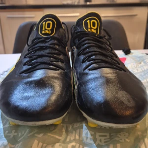 Svarta Puma King fotbollsskor - Säljer ett par svarta Puma King fotbollsskor med gula detaljer och snörning. Skorna har en cool illustration på sidan, gula dobbar och texten 'KING' på plösen och hälen. Tillverkade i skinn för grym känsla på planen.Pele fotbollsskor från Unisport kan diskuteras till lägre pris skriv till mig 