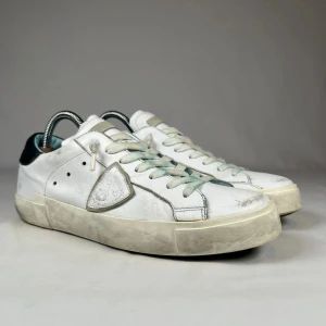  Philippe Model skor - Snygga vita sneakers från Philippe Model med grå detaljer och svart häl. Skorna har en klassisk låg siluett, rund tå och snörning. Tillverkade i skinn med en robust, något smutsad sula för en cool streetkänsla. Perfekta för dig som gillar en avslappnad och trendig look.