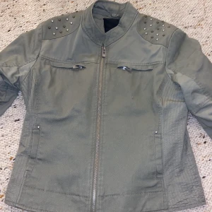 Olivgrön bomberjacka från Amisu - Snygg olivgrön bomberjacka från Amisu med coola nitar i guld och silver på axlarna. Jackan har dragkedja framtill, två bröstfickor med dragkedja och en stilren krage. Perfekt för dig som vill sticka ut med detaljer!