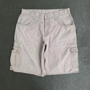 Beige cargo shorts från Interstack - Säljer ett par beige cargo shorts från Interstack med flera fickor och snygga broderade detaljer på ficklocken. Klassisk loose passform och midwaist. Perfekta för chill dagar och streetwear. Materialet är mjuk bomull och de har knappar i trälook.