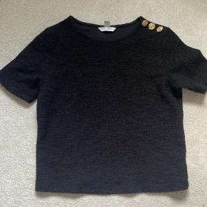 Svart topp med guldknappar från H&M - Svart kortärmad topp från H&M med tre snygga guldknappar på ena axeln🤍
