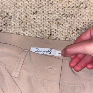 Beige bootcutbyxor från Design by Si - Snygga beige byxor från Design by Si i bootcut-modell. Byxorna har hällor för bälte, två bakfickor och är tillverkade i ett mjukt, lätt material som känns skönt mot huden. Perfekta för dig som gillar en klassisk men trendig look.