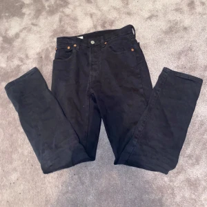 Svarta Levi's 501 jeans - Säljer ett par svarta Levi's 501 jeans i klassisk rak modell. Jeansen är tillverkade i kraftigt denimtyg och har fem fickor, knappgylf och snygga kopparfärgade nitar. Perfekta för dig som gillar tidlös stil och vill ha ett par jeans som funkar till allt. Hör av dig vid minsta fundering! Priset är mutbart.