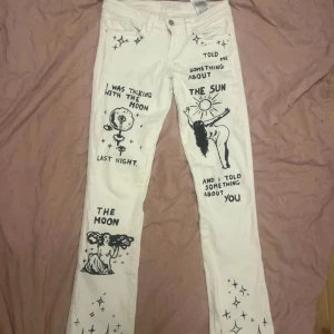 Vita jeans med handmålade motiv - Unika vita jeans med handmålade svarta motiv och texter på benen, samt sol och måne på bakfickorna. Utsvängda passform och klassisk femficksmodell. Perfekta för dig som vill sticka ut med konstnärlig och personlig stil.