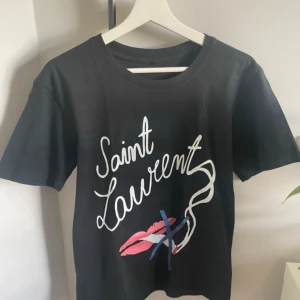 Yves Saint Lauren ”no smoking” t-shirt - Svart t-shirt från Saint Laurent med coolt grafiskt tryck framtill i vitt, rosa och blått. Inga tags Trycket har handskriven text och ett motiv med läppar och linjer. Klassisk rund hals och korta ärmar. Perfekt för dig som gillar statement-plagg och designer.